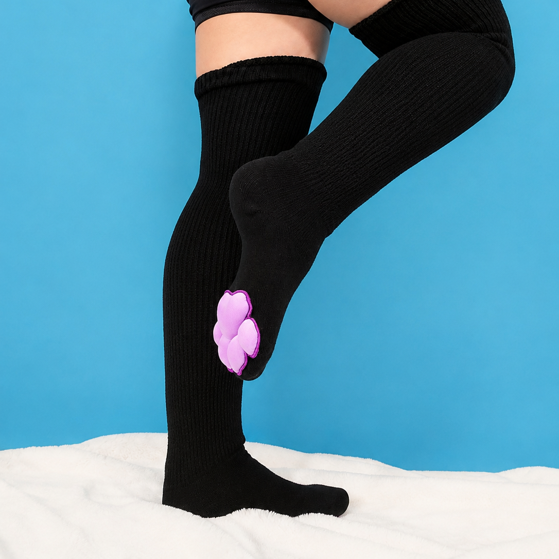 Black ribbed over-the-knee socks worn by a model, featuring a light purple paw-print detail on the lower calf, set against a bright blue background.